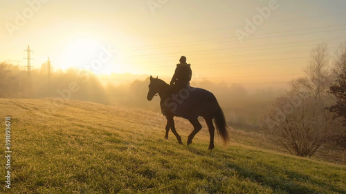 Young woman rides her horse across an empty pasture at a foggy winter sunrise.