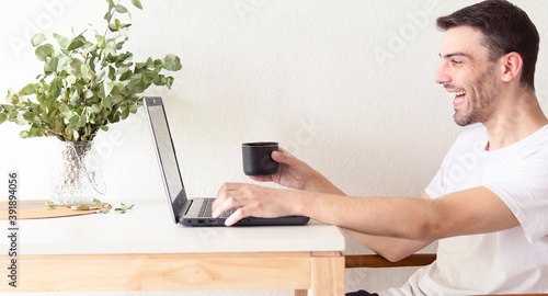 Retrato de un hombre adulto y apuesto caucásico sentado frente a su laptop y bebiendo una taza de café mientras la usa en un apartamento brillante y blanco con luz natural.
