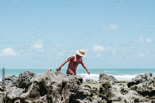 A man walkin down the sun at noon with a hat between the rocks right beside the ocean