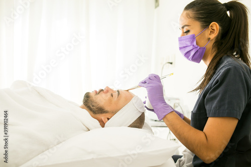 Limpieza facial de un hombre calvo en un spa  con fondo blanco y guantes morados y mujer dermatologa con cubre bocas