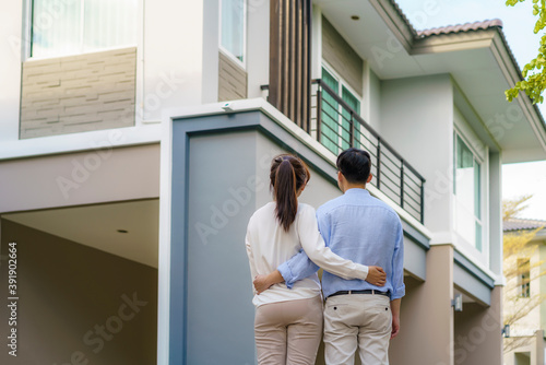 Portrait of Asian young couple standing and hugging together looking happy in front of their new house to start new life. Family, age, home, real estate and people concept.