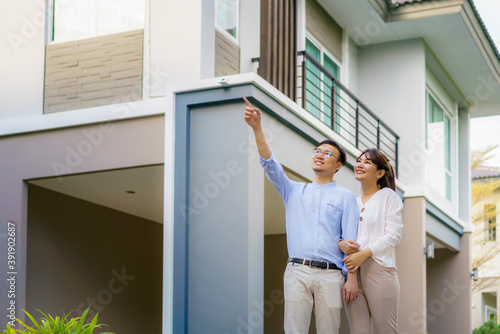 Portrait of Asian couple walking hugging and pointing together looking happy in front of their new house to start new life. Family, age, home, real estate and people concept.