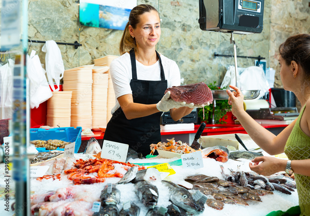 Fine female worker of fish store offering fresh tunny fillet to woman customer