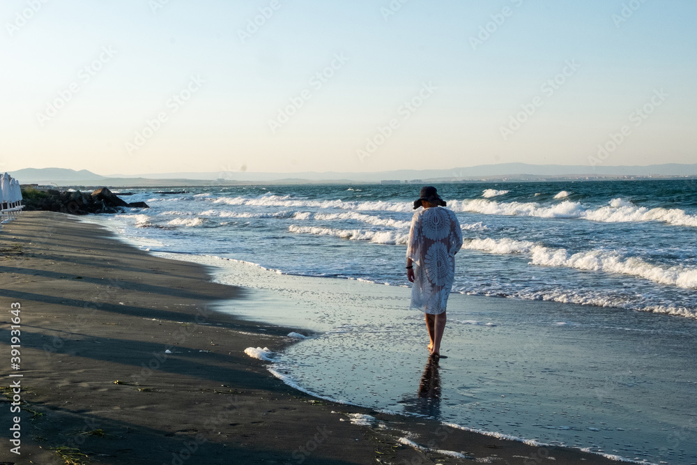 girl walking along the beach. Girl in white long summer dress on vacation. Girl on the shore of the ocean.