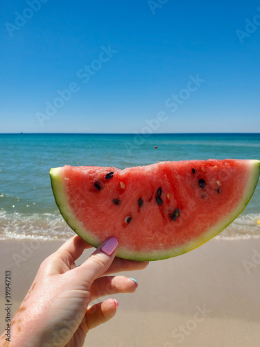one hand holds a slice of fresh and juicy watermelon with a blurred sea and blue sky in summer.