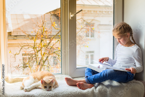 The child is looking at a book while sitting on the windowsill, and her pet is lying nearby. A little girl with a cat is sitting on the windowsill and reading a book.