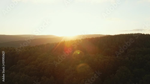 Lens flare in mountains at orange sunset in summer morning aerial view drone. Sun rays. Green pine forest slopes of mountain range. Bright disk of sun in blue sky slowly sets tops of mountains. Nature photo