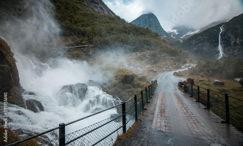wooden bridge over wild waterfall in Norway, epic mountain scenery in the background, moody clouds of fog surrounding the bridge
