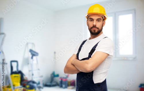 repair, construction and building concept - male worker or builder in yellow helmet and overall with crossed arms over room with equipment on background