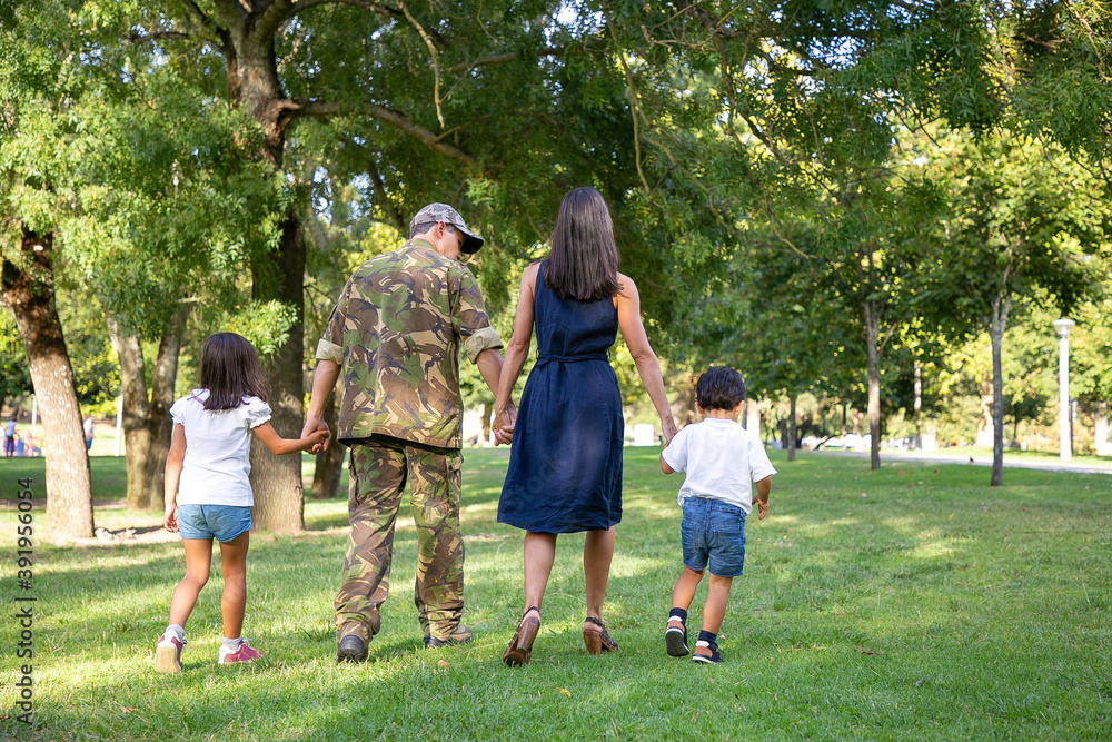 Obraz premium Rear view of Caucasian family holding hands and walking together in city park. Dad in camouflage uniform, long-haired mom and children enjoying holiday on nature. Family reunion and weekend concept