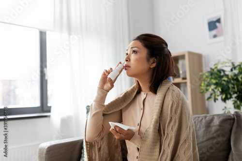 health  cold and people concept - sad sick young asian woman in blanket using nasal spray medicine at home