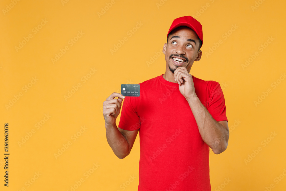 Smiling delivery employee african man 20s in red cap blank print t-shirt uniform workwear work courier dealer service concept hold mockup plastic credit bank card isolated on yellow background studio. Smiling delivery employee african man 20s in red cap blank print t-shirt uniform workwear work courier dealer service concept hold mockup plastic credit bank card isolated on yellow background studio.