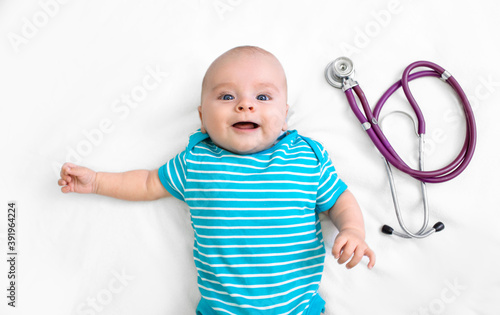 Close-up smiling little baby lies on a white bed near a stethoscope before a pediatrician appointment. Medical caring for a child's health