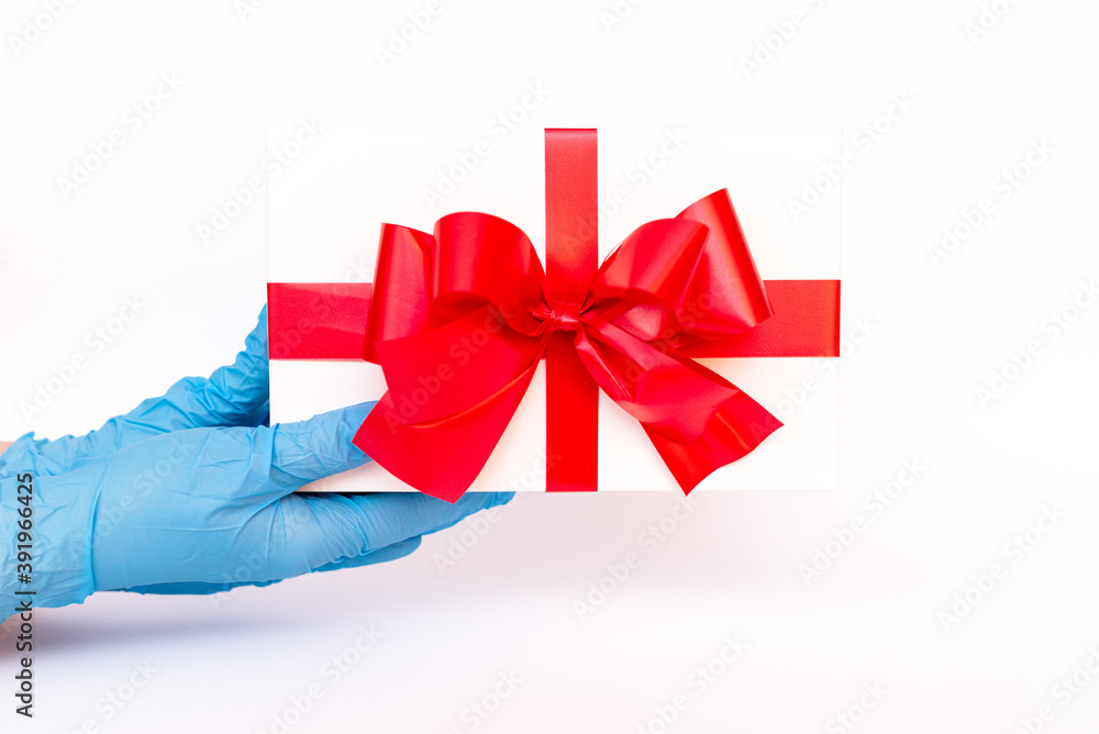Holiday gift.Hands wearing meidical gloves holding gift box with red ribbon.white background.COVID-19 prevention.