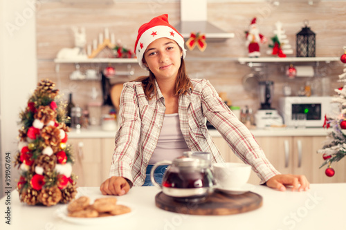 Joyful girl wearing santa heat celebrating christmas in kitchen. Cheerful happy adorable teenager girl in home kitchen with delicious biscuits and xmas tree in the background celebrating winter