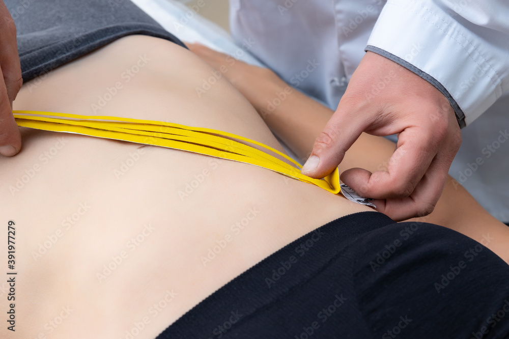 Male physiotherapist applying yellow adhesive elastic therapeutic tape on female abdominal muscle of young adult slim sporty woman lying on couch doctor office. Injury rehabilitation and healthcare - obrazy, fototapety, plakaty 