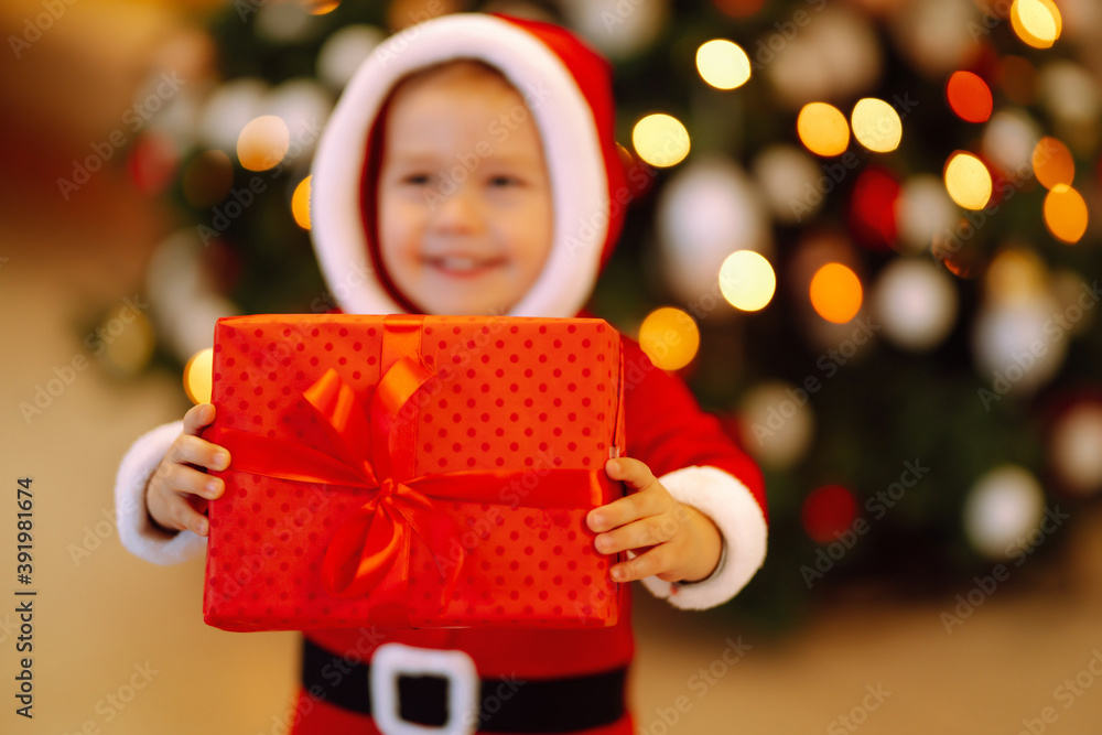Christmas gift in the hand of Little child. Kid in santa costume holding red present box on the background of Christmas lights and tree. Winter holiday, New Year. Christmas gift in the hand of Little child. Kid in santa costume holding red present box on the background of Christmas lights and tree. Winter holiday, New Year.