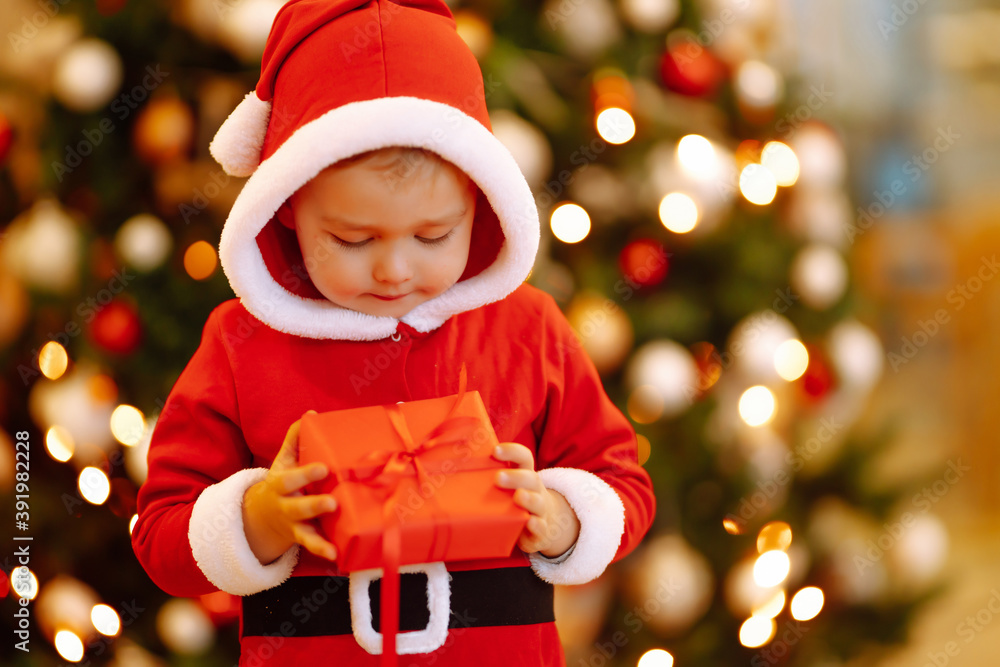 Christmas gift in the hand of Little child.  Kid in santa costume holding red present  box on the background of Christmas lights and tree. Winter holiday, New Year.