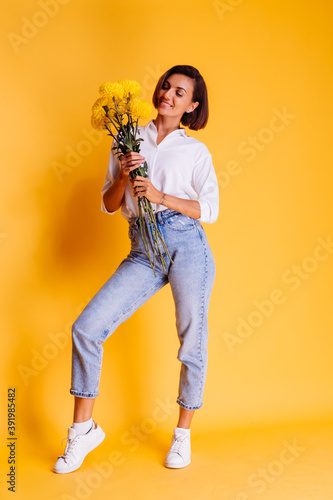 Studio shot on yellow background. Happy caucasian woman short hair wearing casual clothes, white shirt and denim pants, holding bouquet of yellow asters. 