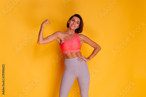 Sport and yoga lifestyle concept. Young fit pretty strong woman dressed in sport clothes, stylish top and leggings, poses against yellow background. Studio shot. 