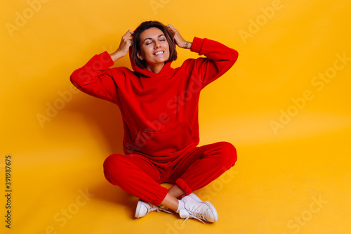 Portrait of happy tanned fit caucasian woman wearing trendy warm red fleece hoodie and pants, getting ready for cold winter. Studio bright shot, yellow background.   © Анастасия Каргаполов
