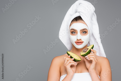 woman with towel on head and clay mask on face holding avocado halves isolated on grey