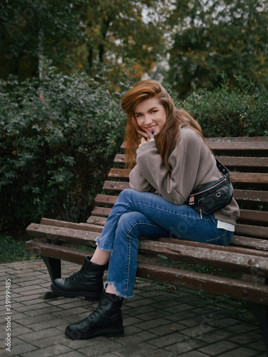 Young thoughtful redhead woman in hoodie hand supports the chin enjoying warm fall day sitting in city park with green and yellow foliage background