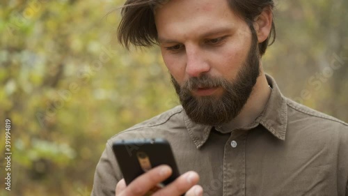 Smiling brutal man in a brown shirt walks in the autumn park holding a phone in his hands and communicates on social networks. Happy person
