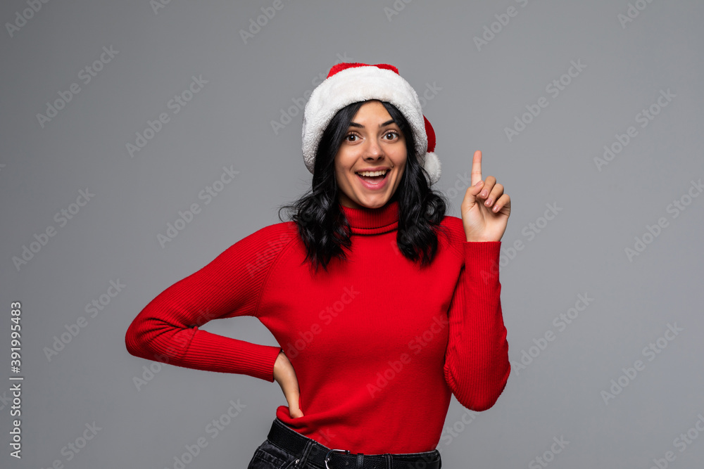 Pretty woman in a red sweater and Santa hat pointing at the copy space up isolated over grey background. Celebrating Christmas and New Year Pretty woman in a red sweater and Santa hat pointing at the copy space up isolated over grey background. Celebrating Christmas and New Year