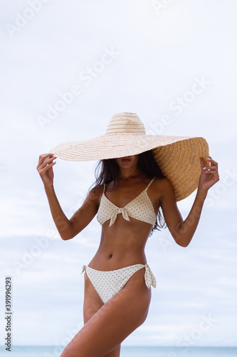 Fashion portrait of fit tanned slim stylish woman wearing large big giant straw hat on rocky tropical beach, natural warm light. 