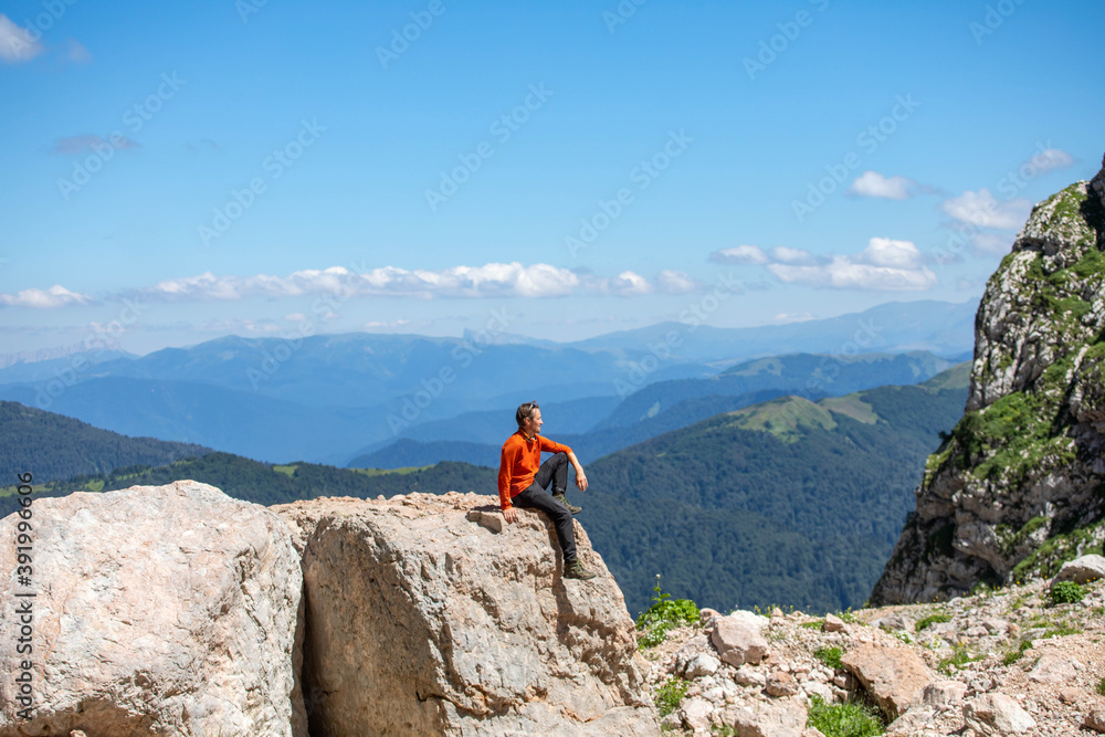 A man in an orange sweater sits on a stone against a background of high mountains