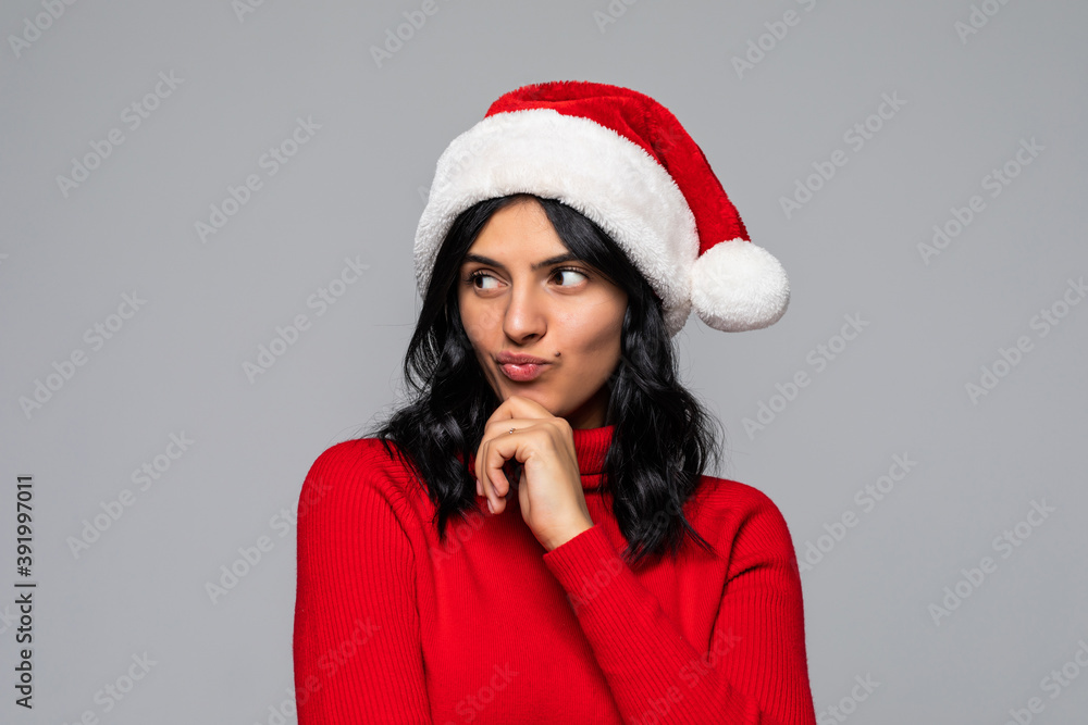 Portrait of a cute christmas woman with a red santa claus hat, finger on chin, confused, thinking what to shop for the holiday season. Emotion on isolated grey background. Portrait of a cute christmas woman with a red santa claus hat, finger on chin, confused, thinking what to shop for the holiday season. Emotion on isolated grey background.