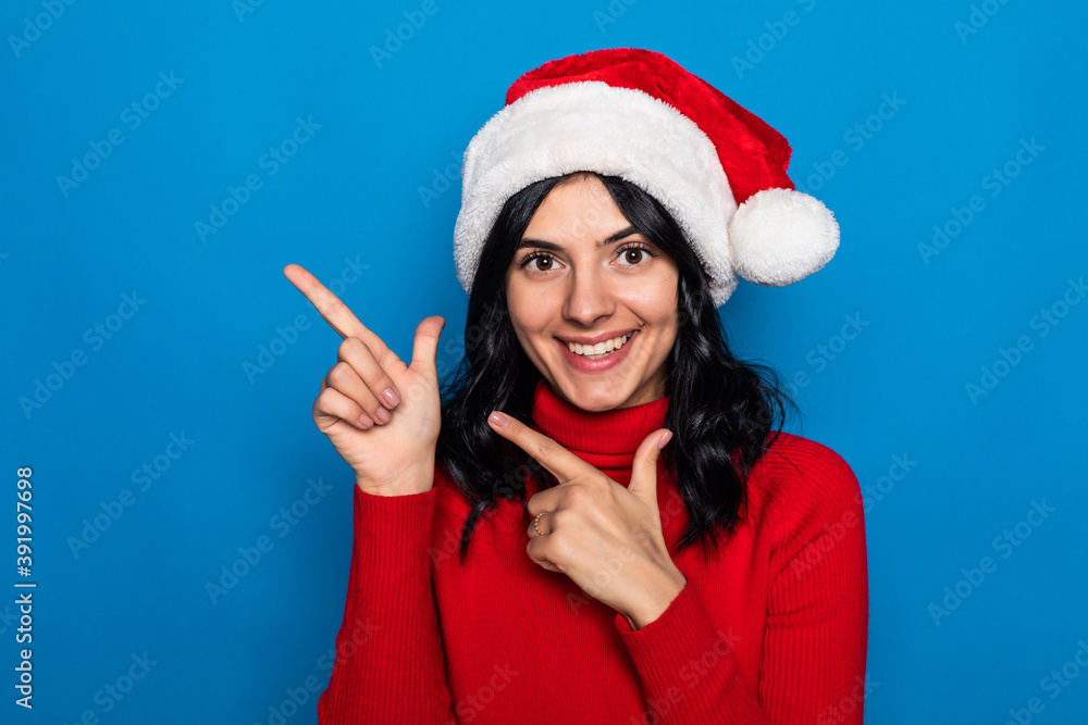 Woman with christmas hat pointing to the side to present a product isolated on blue background Woman with christmas hat pointing to the side to present a product isolated on blue background