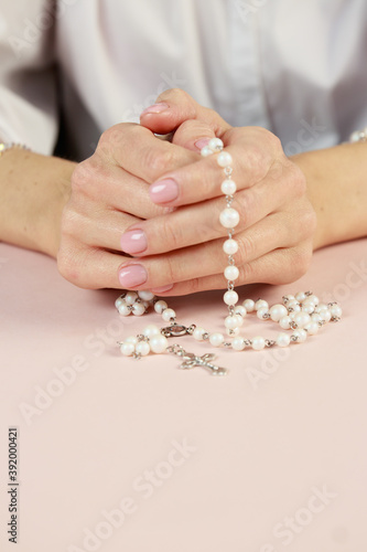 Hands with white rosary folded in prayer. Beige delicate background.