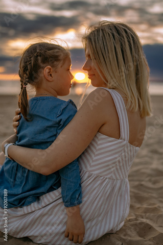 young blond caucasian woman sitting with her daughter on beach looking at sunset. Time together. Family concept. Image with selective focus © Yulia Raneva