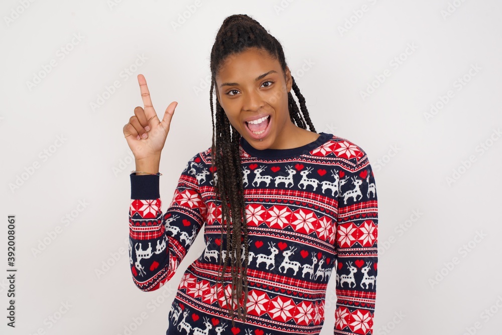 Pleasant looking Young beautiful African American woman wearing Christmas sweater against white wall has clever expression, raises one finger, remembers himself not to forget tell important thing. Pleasant looking Young beautiful African American woman wearing Christmas sweater against white wall has clever expression, raises one finger, remembers himself not to forget tell important thing.