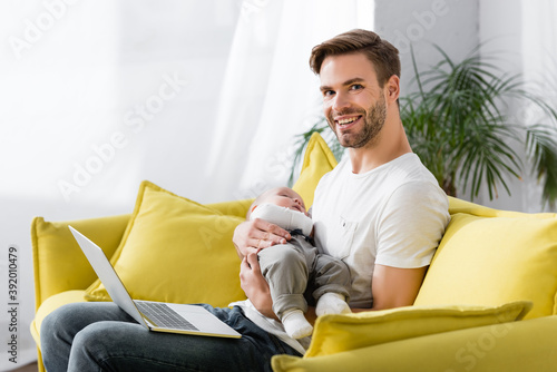 happy father holding in arms sleepy baby son while sitting on sofa with laptop