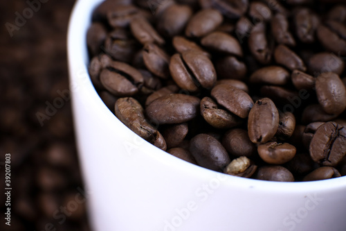 White cup full of coffee beans close up against the coffee beans background. Coffee mug. Morning espresso.