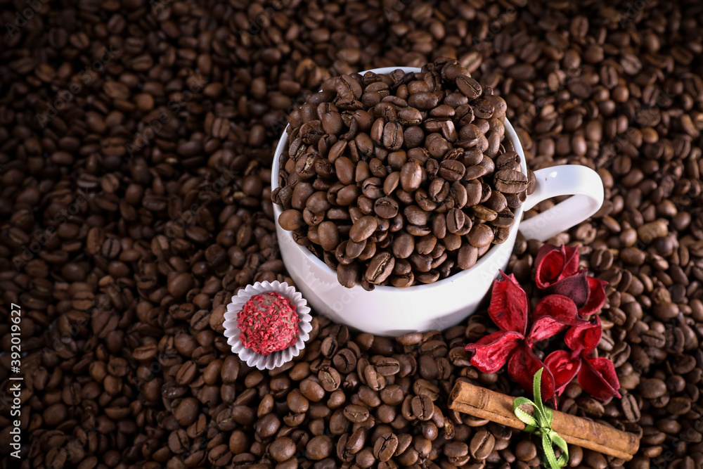 White cup full of coffee beans on a coffee beans background with red flowers , red chocolate and cinnamon. Morning espresso. Coffee mug. White cup full of coffee beans on a coffee beans background with red flowers , red chocolate and cinnamon. Morning espresso. Coffee mug.