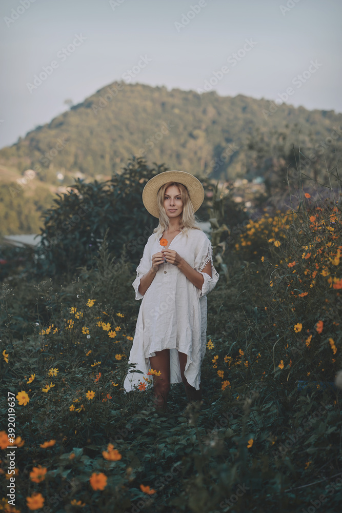 The blonde in a linen dress and straw hat walks among herbs and wildflowers on a meadow in the mountains. A woman in the Boho style, resting in the countryside, a simple village lifestyle