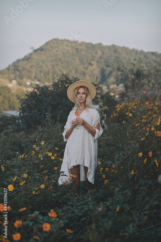Obraz na płótnie The blonde in a linen dress and straw hat walks among herbs and wildflowers on a meadow in the mountains. A woman in the Boho style, resting in the countryside, a simple village lifestyle w salonie