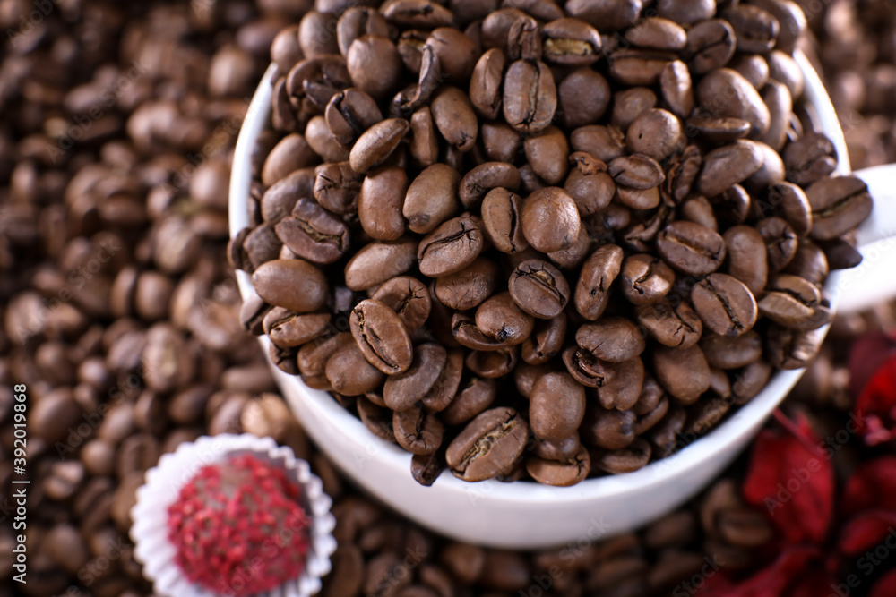 Fototapeta premium White cup full of coffee beans on a coffee beans background with red chocolate candy and red flowers. Morning espresso. Coffee mug.