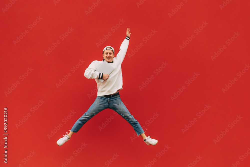 Happy guy in stylish casual clothes jumps on a background of a red wall with a smile on his face and a laptop in his hands. Background. Isolated.