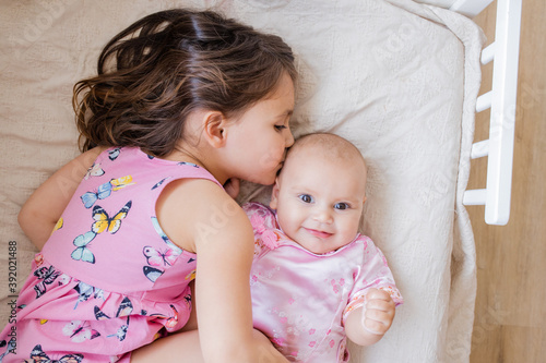 Little girl lovingly hugging and kissing her happy baby sister on a bed