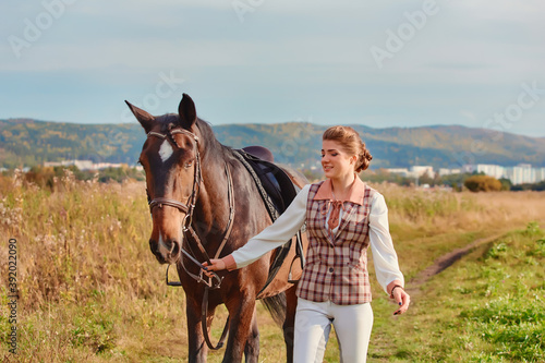 A beautiful slender girl in a sports rider costume leads her horse across a meadow.