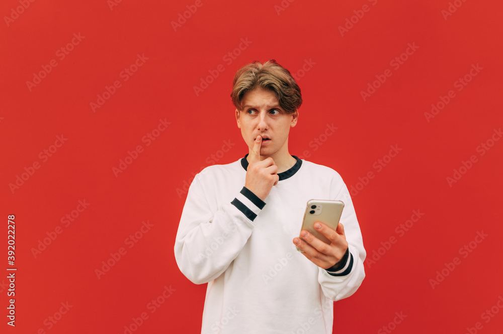 Pensive young man stands on a red background with a smartphone in his hand and looks thoughtfully to the side on copy space. Isolated on red wall background. Pensive young man stands on a red background with a smartphone in his hand and looks thoughtfully to the side on copy space. Isolated on red wall background.