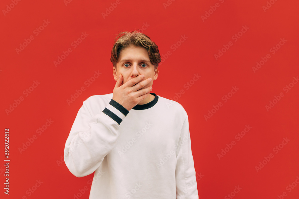 Shocked young man covers his face with his hand and looks at the camera with a shocked face on a background of red wall. Isolated. Shocked young man covers his face with his hand and looks at the camera with a shocked face on a background of red wall. Isolated.