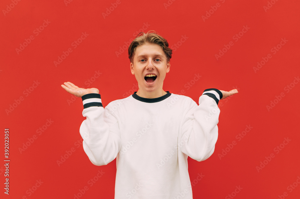 positive guy on a red background, spreads his arms to the side and looks at the camera with a smile on his face. Young man with a smile on his face does not know the answer, shows a gesture positive guy on a red background, spreads his arms to the side and looks at the camera with a smile on his face. Young man with a smile on his face does not know the answer, shows a gesture