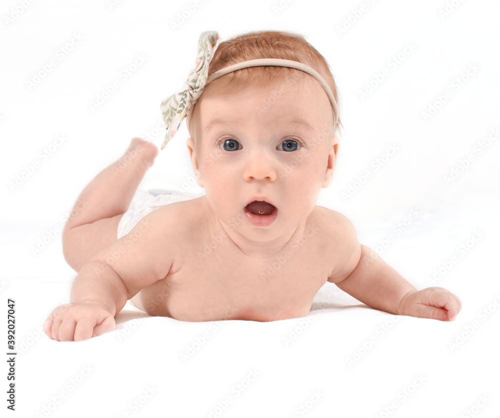 Adorable, amazed, caucasian baby girl lying on stomach, on white blanket with bow on her head, looking at camera, on white background. Open mouth, healthy skin. Baby care concept. Adorable, amazed, caucasian baby girl lying on stomach, on white blanket with bow on her head, looking at camera, on white background. Open mouth, healthy skin. Baby care concept.