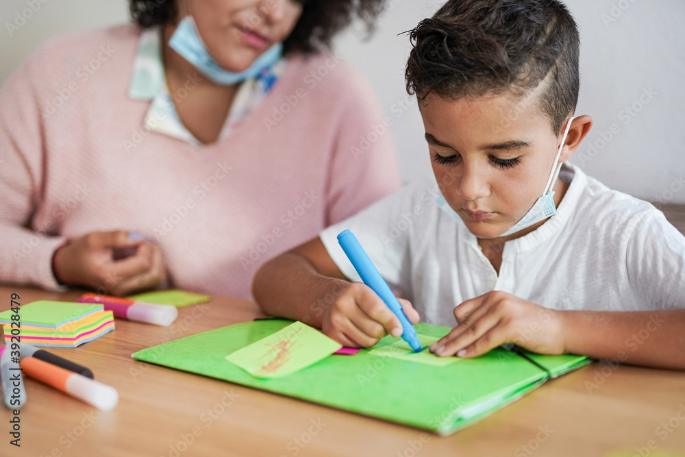 Little boy draws at school while wearing surgical face mask under chin - Teacher in the background Little boy draws at school while wearing surgical face mask under chin - Teacher in the background